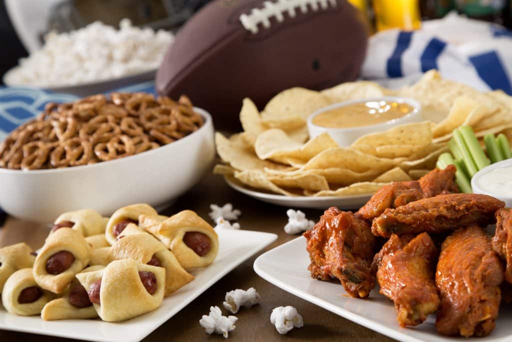 football tailgating spread with pretzels, nachos, hot wings, and pigs in a blanket